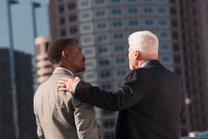 Ein älterer Mann legt einem jüngeren Mann im Business-Kontext die Hand auf die Schulter – Symbol für Generationswechsel und Unternehmensnachfolge.